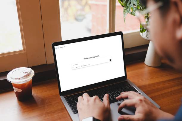 Person using a laptop with the screen displaying a chat interface, sitting by a window with a coffee on the desk.