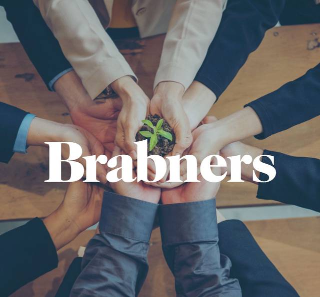 Overhead view of business-attired people's hands cupped around a green seedling in soil on a wooden table, with the white word Brabners overlaid