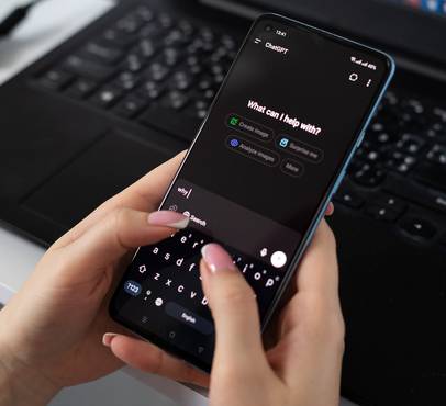 A person's hands hold a smartphone open to the ChatGPT app in dark mode, typing "why" with a pink-nailed finger, with a laptop keyboard and earphones on the desk behind.