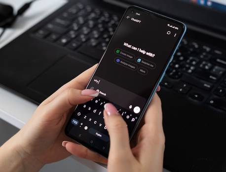 A person's hands hold a smartphone open to the ChatGPT app in dark mode, typing "why" with a pink-nailed finger, with a laptop keyboard and earphones on the desk behind.
