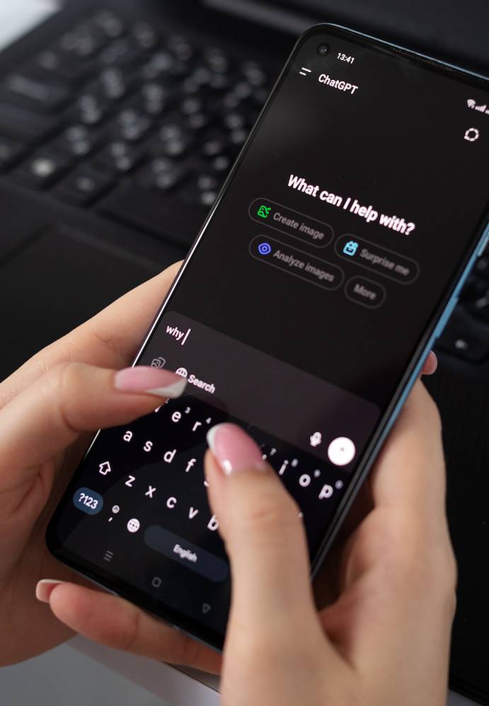 A person's hands hold a smartphone open to the ChatGPT app in dark mode, typing "why" with a pink-nailed finger, with a laptop keyboard and earphones on the desk behind.