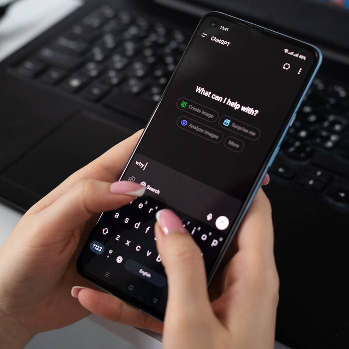 Person holding a smartphone with ChatGPT app open, keyboard and laptop in the background on a desk.