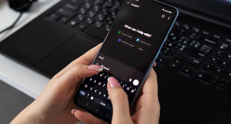 Person holding a smartphone with ChatGPT app open, keyboard and laptop in the background on a desk.