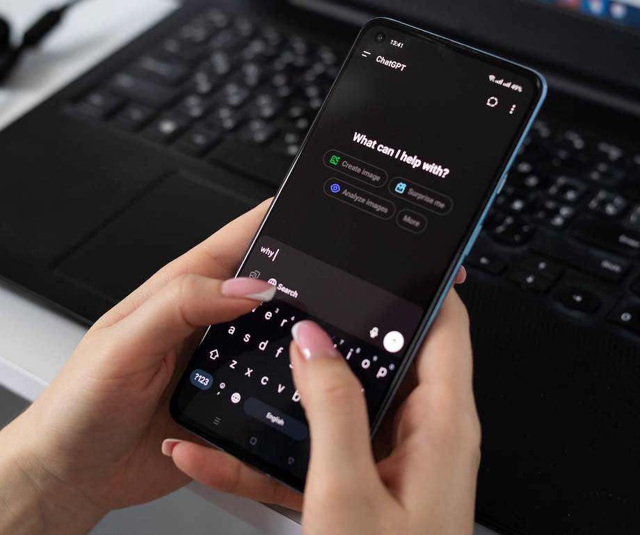A person's hands hold a smartphone open to the ChatGPT app in dark mode, typing "why" with a pink-nailed finger, with a laptop keyboard and earphones on the desk behind.