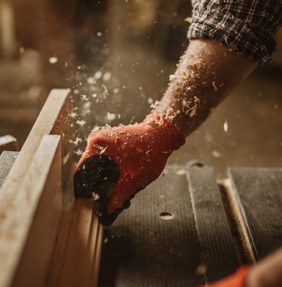 A person using a saw with a red glove to cut a piece of wood, with sawdust flying in the air and a woodwork workshop background.