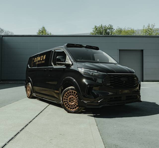 Black customised van with gold accents and "Demon Conversions" branding, parked outside a grey industrial building with roof-mounted equipment visible.