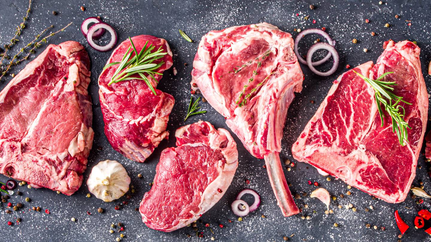 Various raw beef cuts, seasoned with herbs, garlic, and pepper, arranged on a dark surface with scattered spices and onion rings.