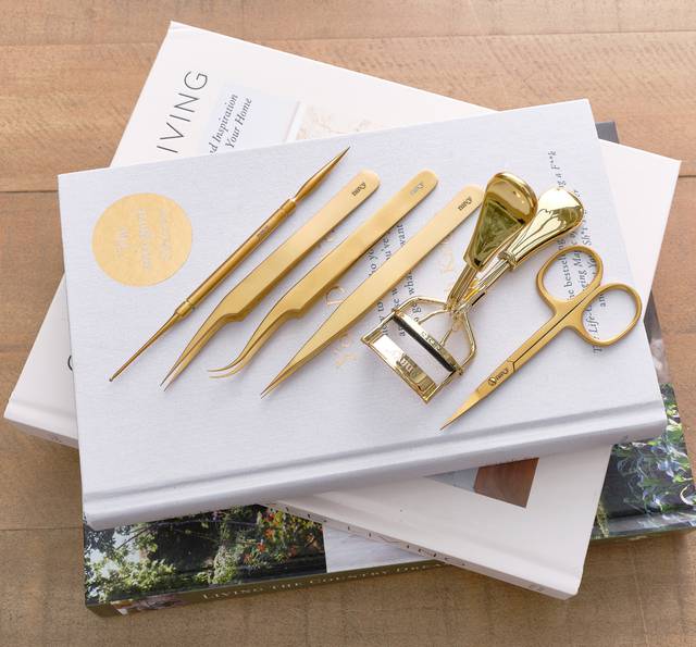 Set of gold-toned grooming tools and accessories arranged on a white hardcover book, including scissors, tweezers, a nail file set, and a nail cutter, atop magazines.