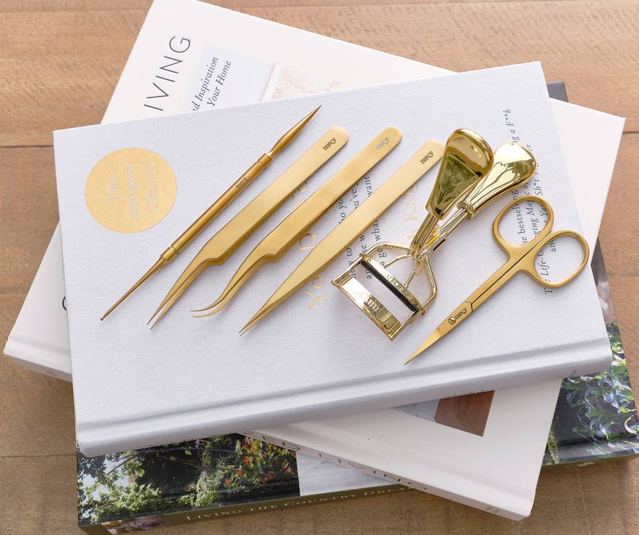 Set of gold-toned grooming tools and accessories arranged on a white hardcover book, including scissors, tweezers, a nail file set, and a nail cutter, atop magazines.