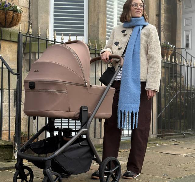 Person with glasses wearing a cream coat and blue scarf standing with a pink pram on a city street lined with stone buildings and an iron fence.