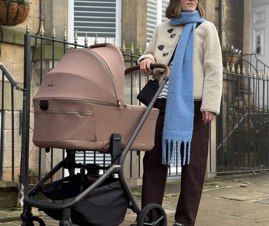 Person with glasses wearing a cream coat and blue scarf standing with a pink pram on a city street lined with stone buildings and an iron fence.