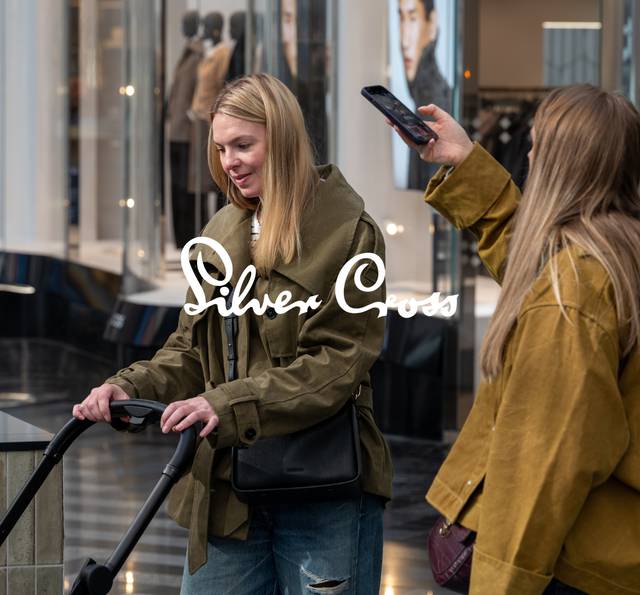 Two people shopping in a mall, one holding an electric scooter and the other taking a photo with a smartphone, with "Silver Cross" text overlay.