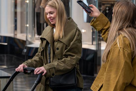 Two people in a modern shopping mall corridor, one holding an electric scooter and the other taking a photo with a smartphone, both wearing autumn jackets.