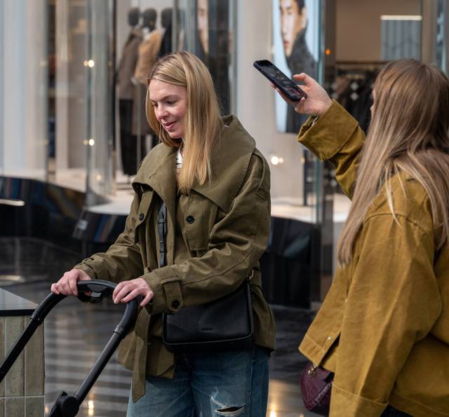 Two people in a modern shopping mall corridor, one holding an electric scooter and the other taking a photo with a smartphone, both wearing autumn jackets.