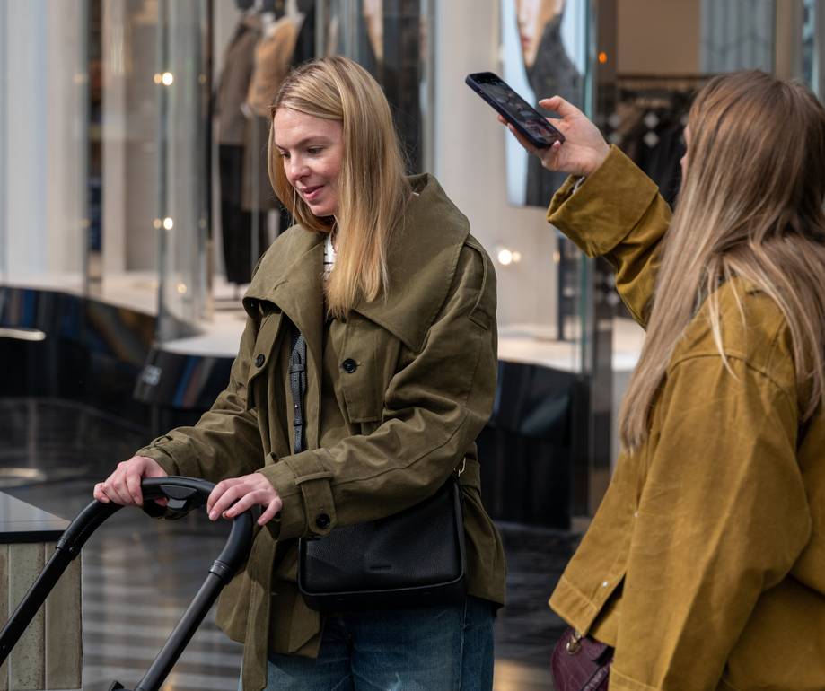 Two people in a modern shopping mall corridor, one holding an electric scooter and the other taking a photo with a smartphone, both wearing autumn jackets.