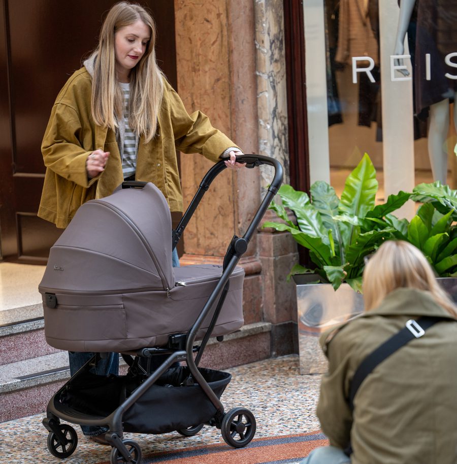 Young person in mustard jacket pushing a grey baby pram on a city street, with another person visible in foreground wearing olive jacket, near shop storefronts and potted plants.