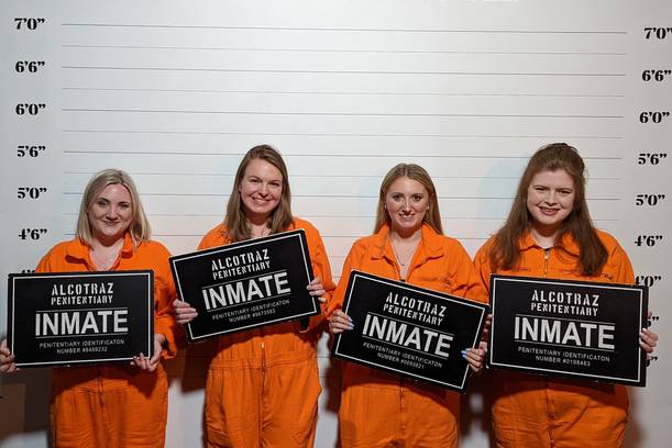 Four women in orange prison jumpsuits holding black and white inmate signs stand in front of a height chart background reading "ALCOTRAZ".