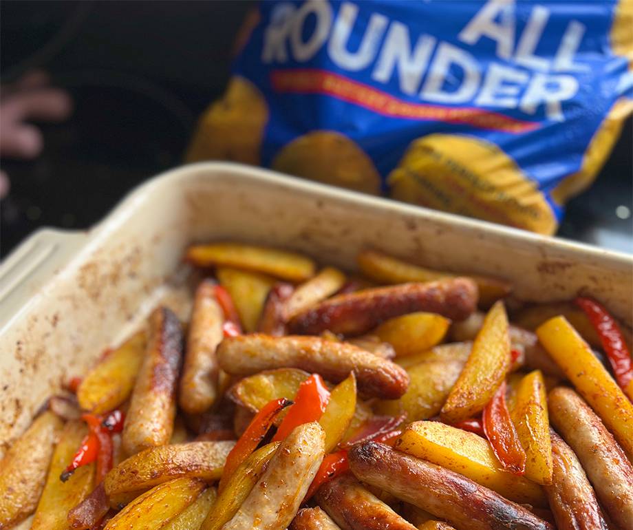 Golden roasted potato wedges, sausages and red pepper strips in a baking dish, with a blue All Rounder potato bag behind.