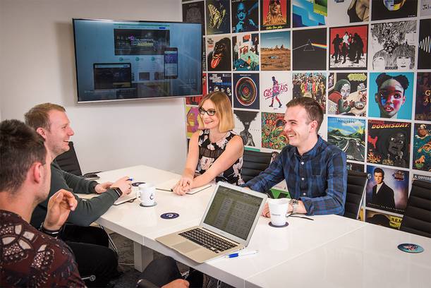 Four young professionals sit around a white conference table, smiling and talking, with a colourful wall of posters behind them.