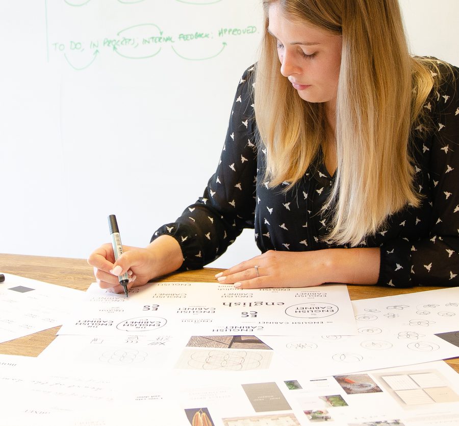 A woman with long blonde hair is seated at a desk, writing on a large sheet of paper with various sketches and notes, with a whiteboard in the background.