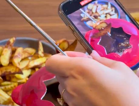 A person taking a photo of potato wedges with a smartphone, with a bowl of wedges in the background on a wooden surface.
