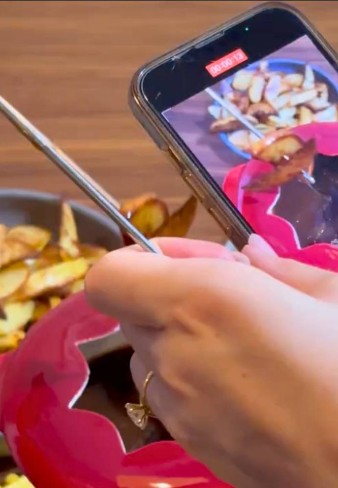 A person taking a photo of potato wedges with a smartphone, with a bowl of wedges in the background on a wooden surface.
