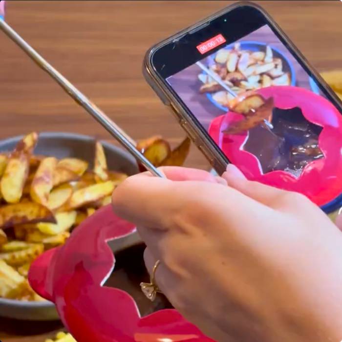 A person taking a photo of potato wedges with a smartphone, with a bowl of wedges in the background on a wooden surface.
