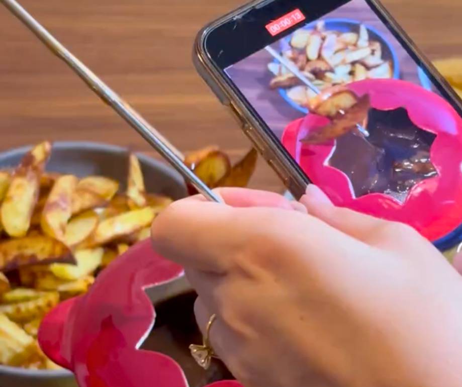 A person taking a photo of potato wedges with a smartphone, with a bowl of wedges in the background on a wooden surface.