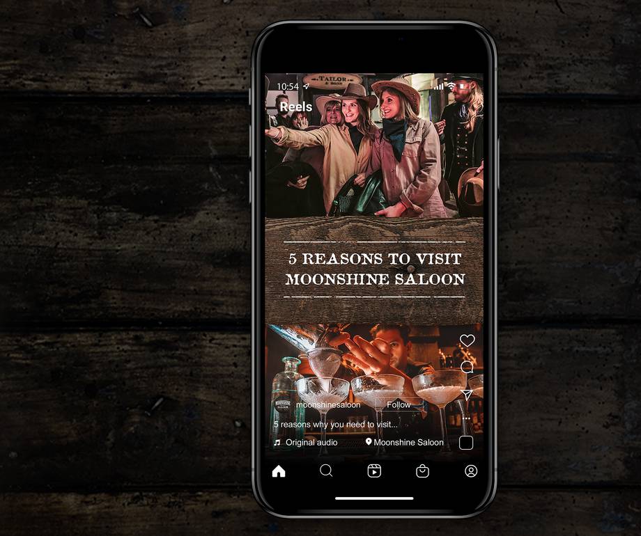 A smartphone displaying a social media profile for Moonshine Saloon, featuring photos of people in western attire and cocktails, on a dark wooden surface.