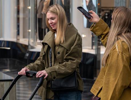Two women, with one pushing a trolley and the other taking a photo, are standing inside a shopping mall with mannequins displayed in the background.