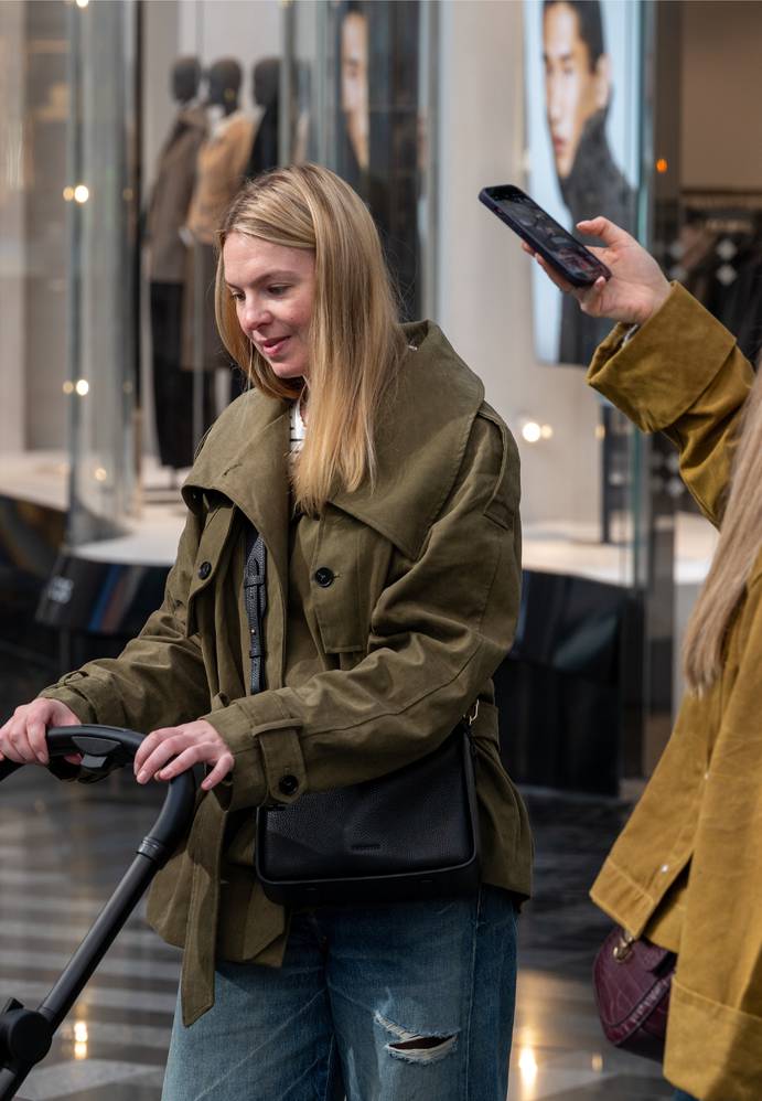 Two women, with one pushing a trolley and the other taking a photo, are standing inside a shopping mall with mannequins displayed in the background.