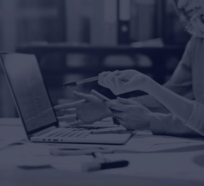 Two people working together, one using a tablet and the other holding a pen, seated at a desk with a laptop and stationery in an office setting.