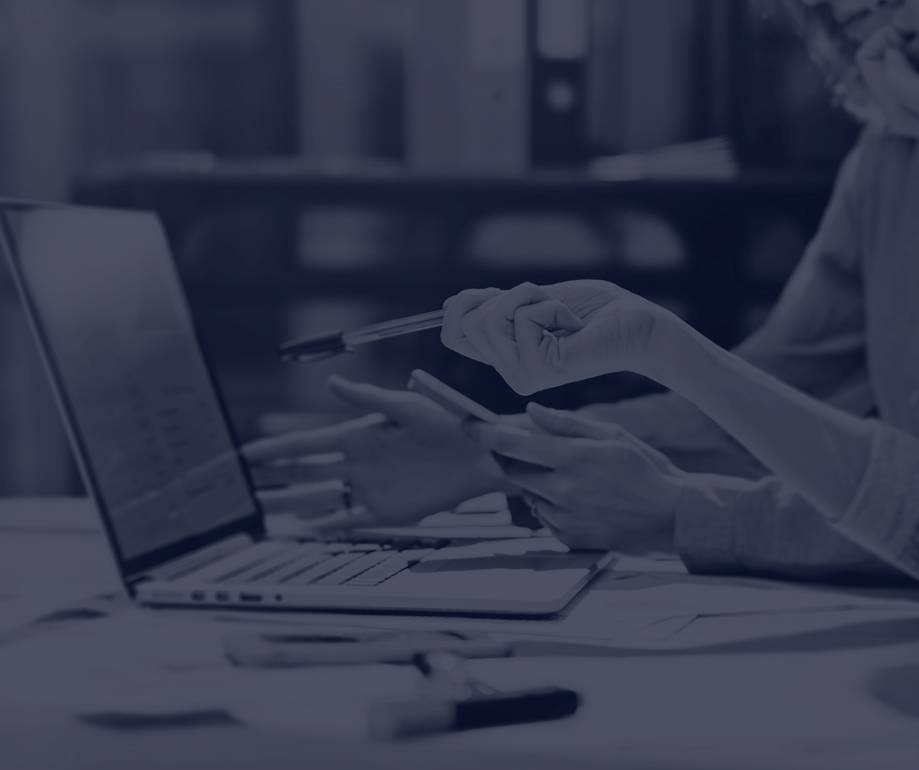 Two people working together, one using a tablet and the other holding a pen, seated at a desk with a laptop and stationery in an office setting.
