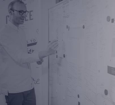 A man with glasses writing on a large whiteboard covered in sketches and notes, standing next to various diagrams and patterns in a creative workspace.
