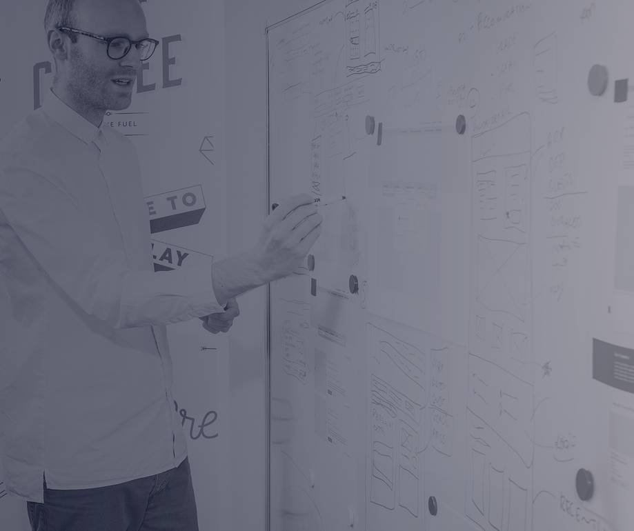 A man with glasses writing on a large whiteboard covered in sketches and notes, standing next to various diagrams and patterns in a creative workspace.