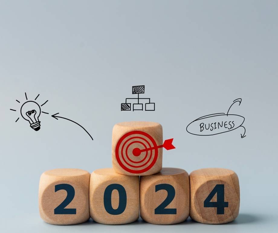 Wooden blocks display the year 2024 with the '0' block featuring a red target icon, surrounded by sketches of a lightbulb, organisational chart, and "Business".