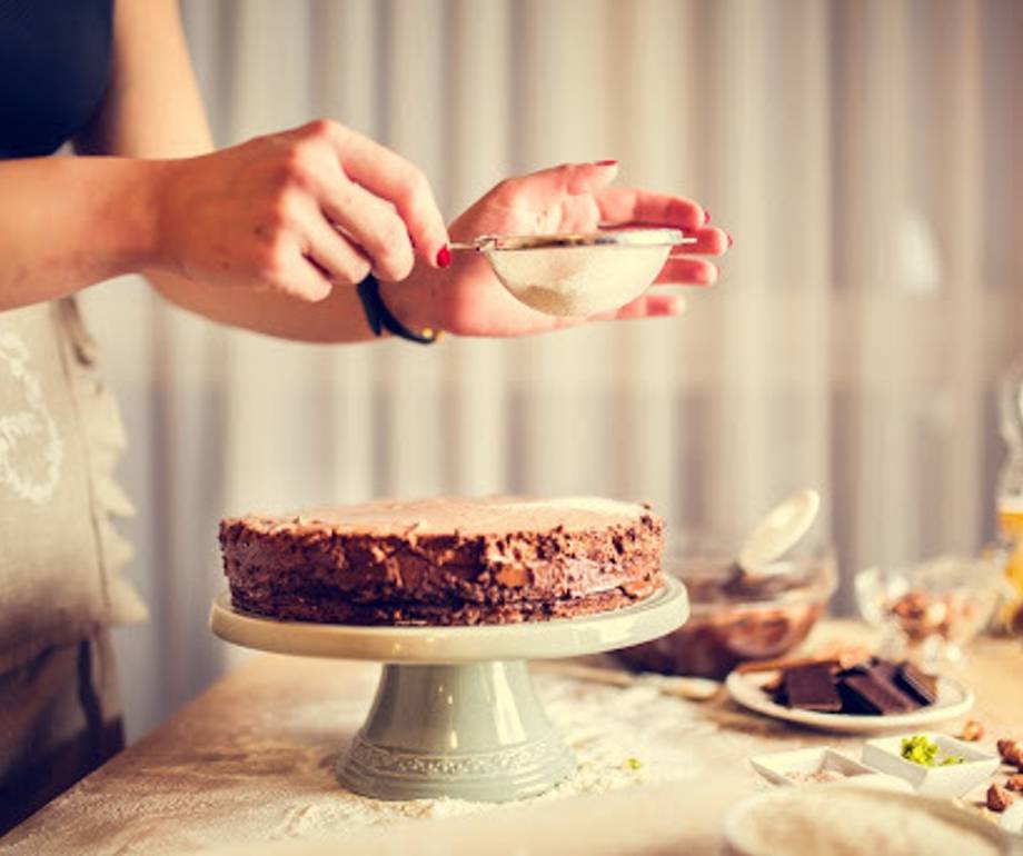 Close-up of a person decorating a cake with a piping bag, with various dessert ingredients and tools on the table in the background.