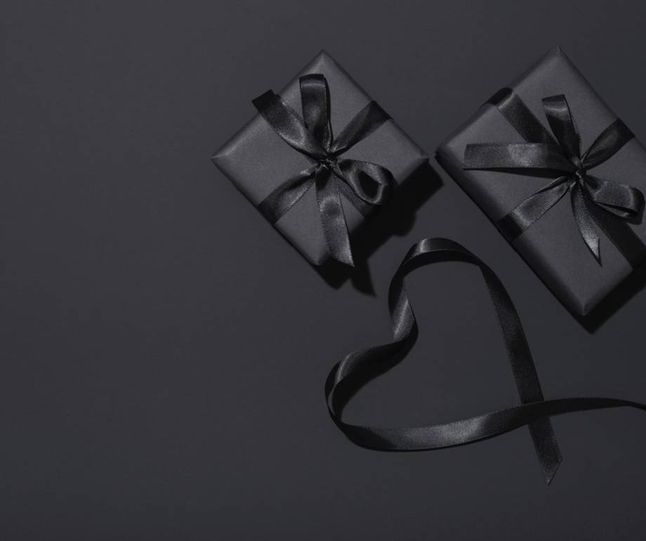 Two black gift boxes with black satin ribbon bows on a dark grey background, with a looped ribbon curling into a heart shape below.