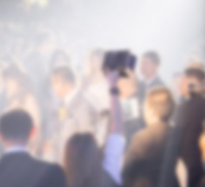 A blurred crowd of people at an event, with some standing and others sitting, and a person holding a camera or phone in the centre.
