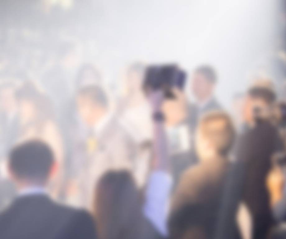 A blurred crowd of people at an event, with some standing and others sitting, and a person holding a camera or phone in the centre.