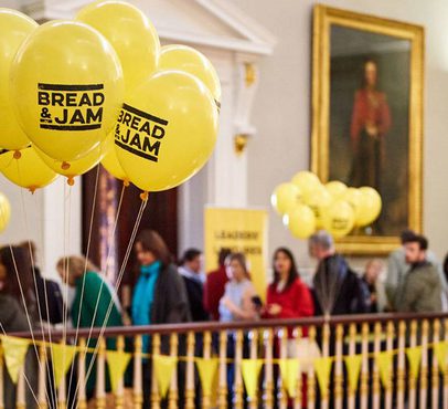 Yellow balloons with "BREAD & JAM" written on them, floating above a crowd in an ornate room with golden accents and portrait paintings.