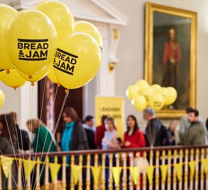 Yellow balloons with "BREAD & JAM" written on them, floating above a crowd in an ornate room with golden accents and portrait paintings.
