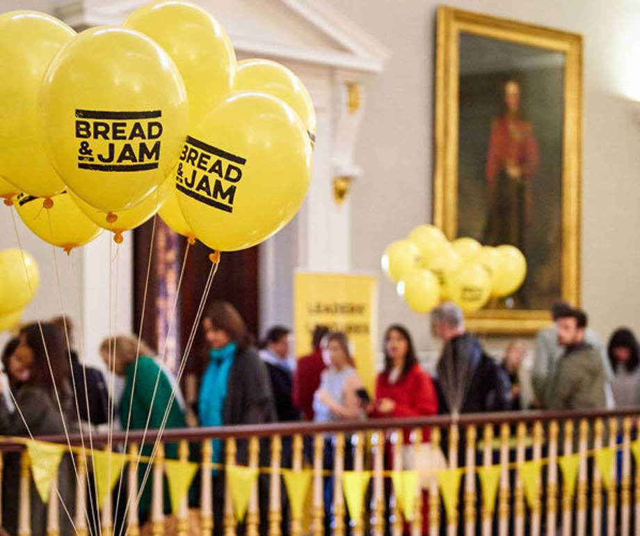 Yellow balloons with "BREAD & JAM" written on them, floating above a crowd in an ornate room with golden accents and portrait paintings.