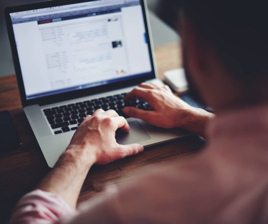 Person using a laptop at a wooden desk, viewing a social media profile or webpage on the screen.