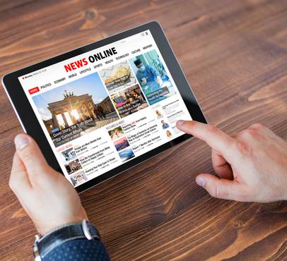 Person holding a tablet displaying a news website, with a wooden table surface in the background and the person wearing a dark blue shirt.