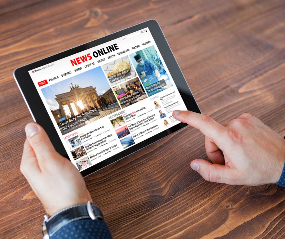 Person holding a tablet displaying a news website, with a wooden table surface in the background and the person wearing a dark blue shirt.