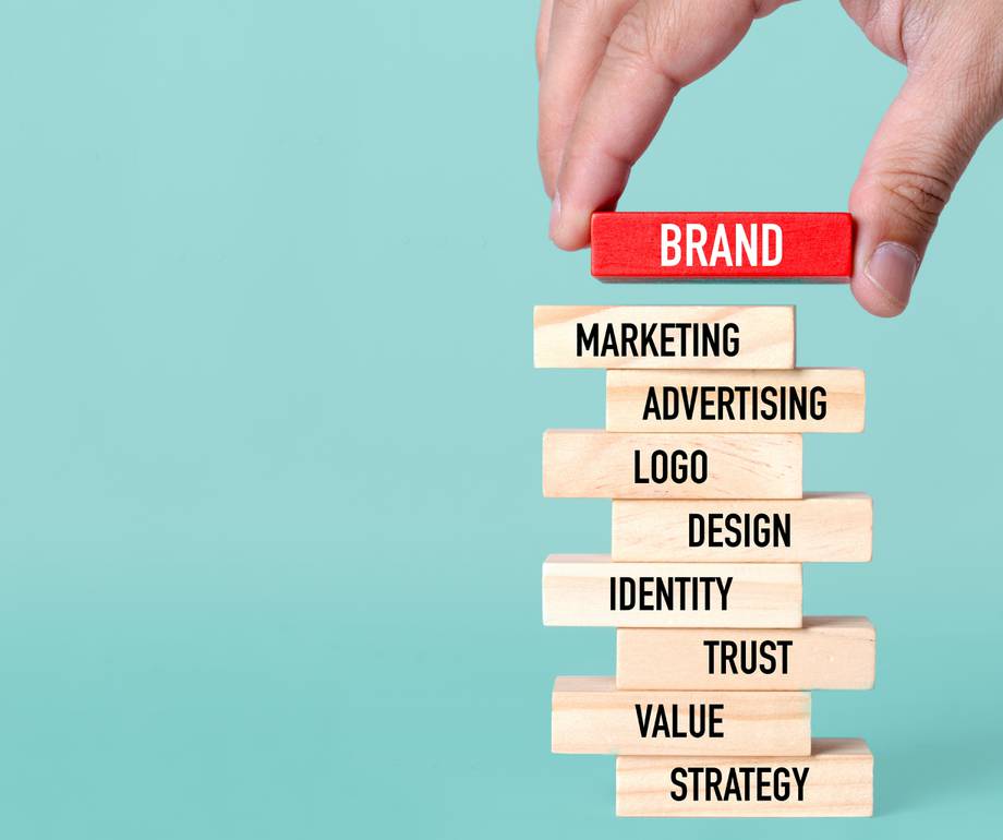 Hand placing a red block labeled "BRAND" on top of stacked wooden blocks with words related to marketing and branding on a light blue background.