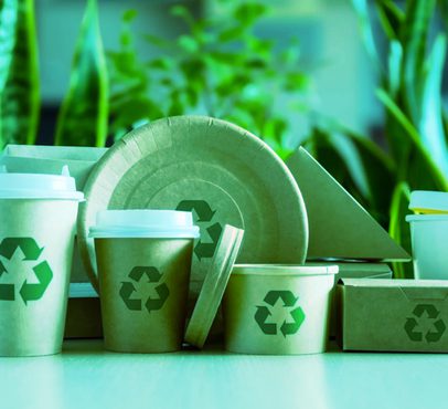 Recyclable coffee cups and packaging with recycling symbols on a table, greenery in the background, emphasising eco-friendly packaging.