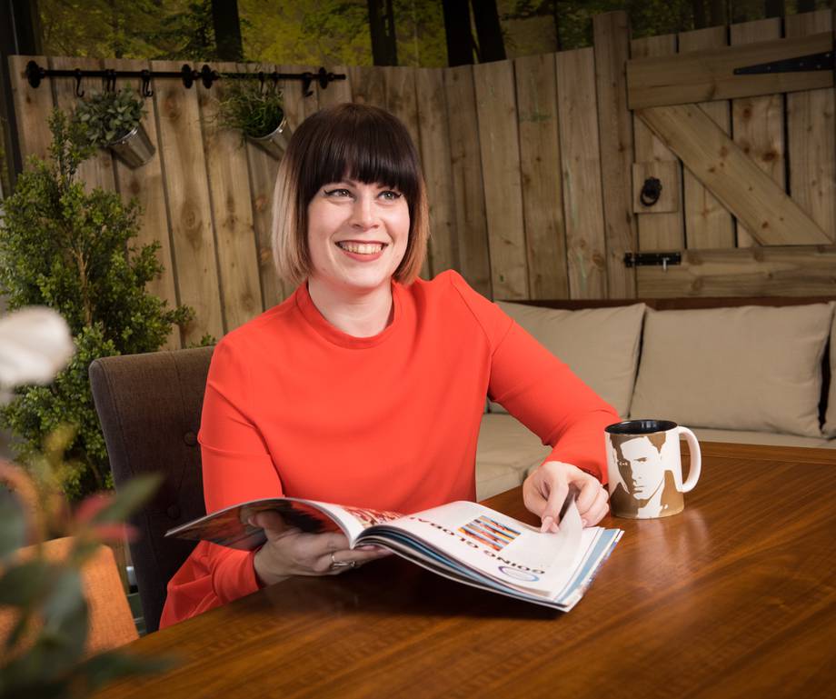 Woman with dark hair and bangs in a red top, sitting at a wooden table with a magazine and a black-and-white mug, smiling in a cozy outdoor setting.
