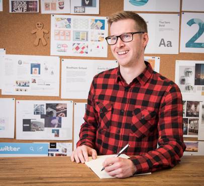 A smiling man with glasses in a red and black checkered shirt sitting at a desk with a notebook, in front of a bulletin board covered with design posters.